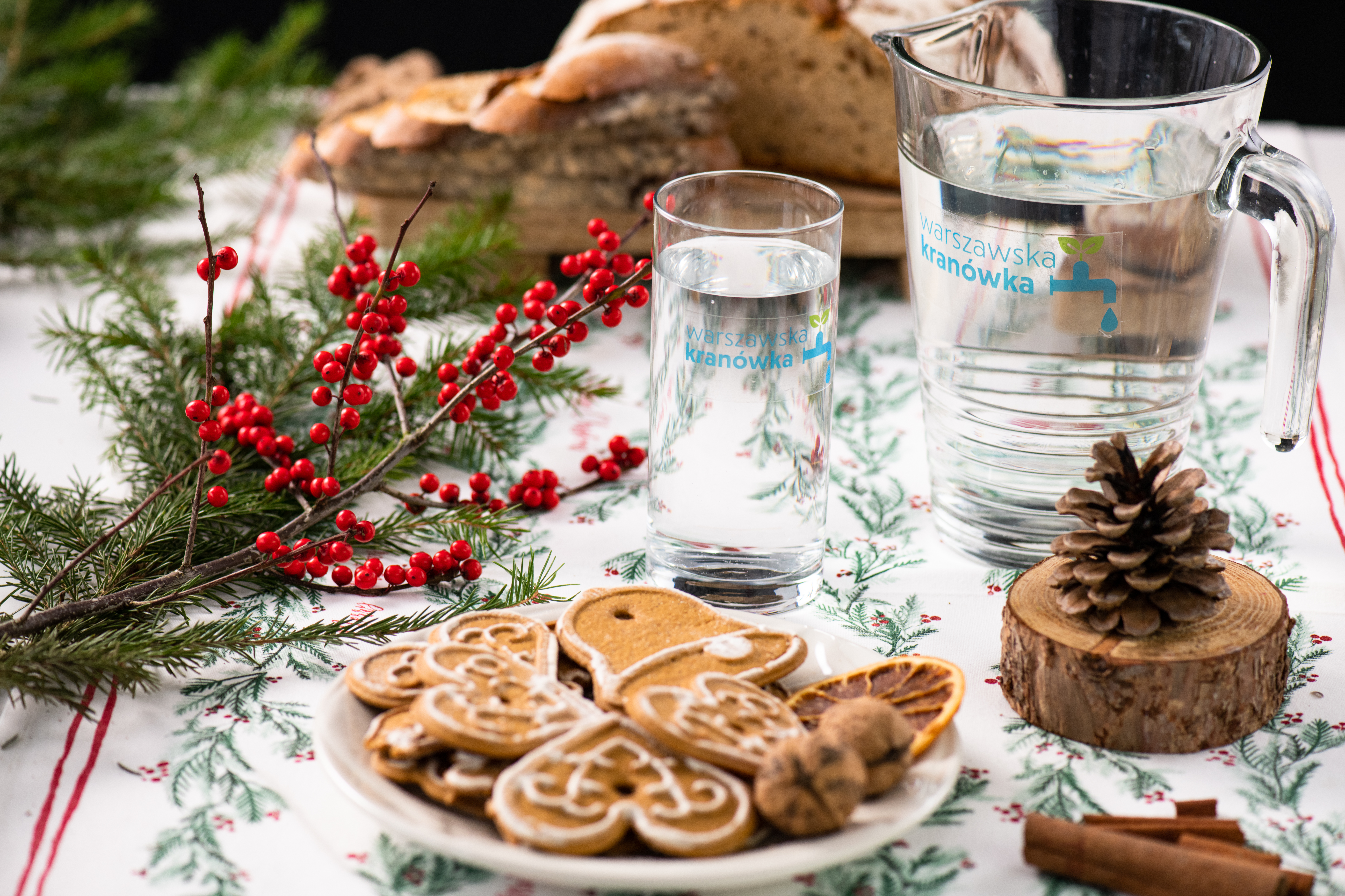 Stół z obrusem, na nim pierniki, dekoracja świąteczna, chleb oraz dzbanek i szklanka z wodą.