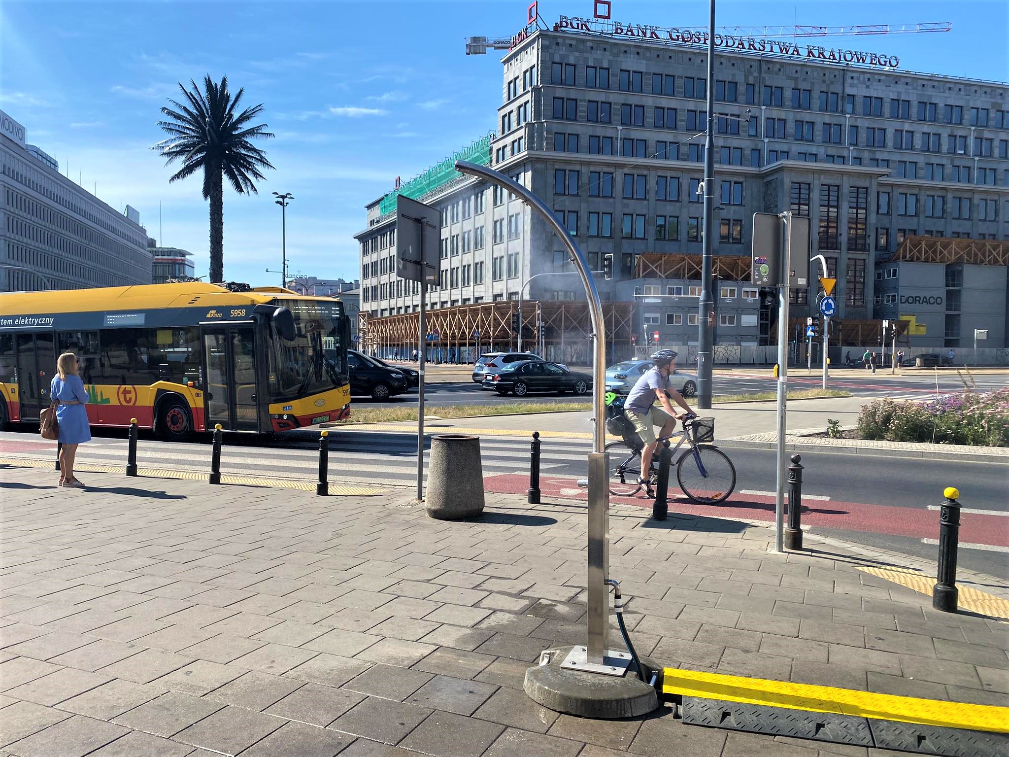 Urządzenie w kształcie luku na tle miasta. W tle budynek wielopiętrowy, palma, po lewej autobus miejski.