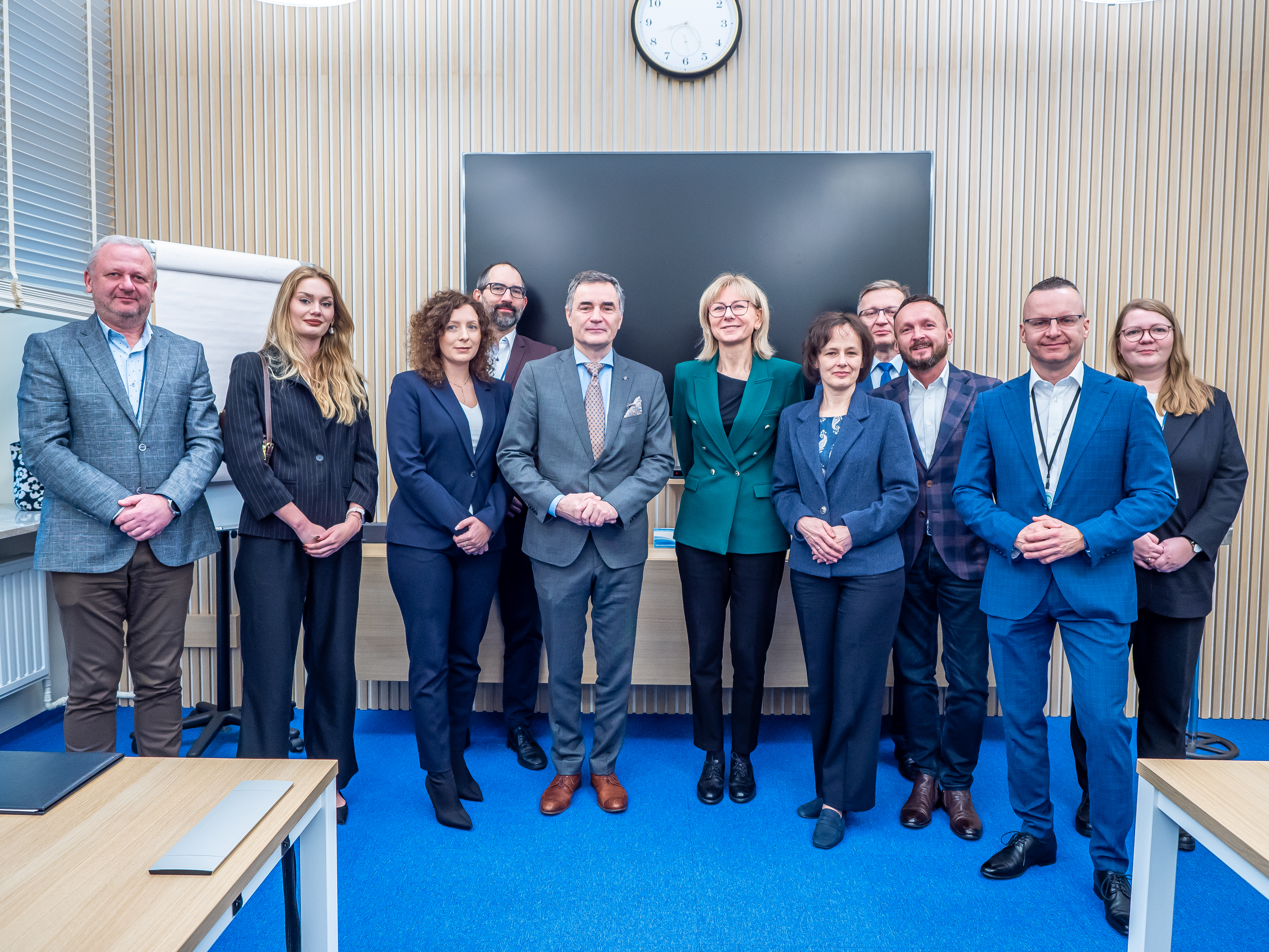 Prezesi Renata Tomusiak i Pawel Orlof ściskaja sobie ręce, usmiechają się. Pani prezes trzyma w ręce tecjzke z dokumentami. W tle na ścianie tapeta z panoramą Warszawy.