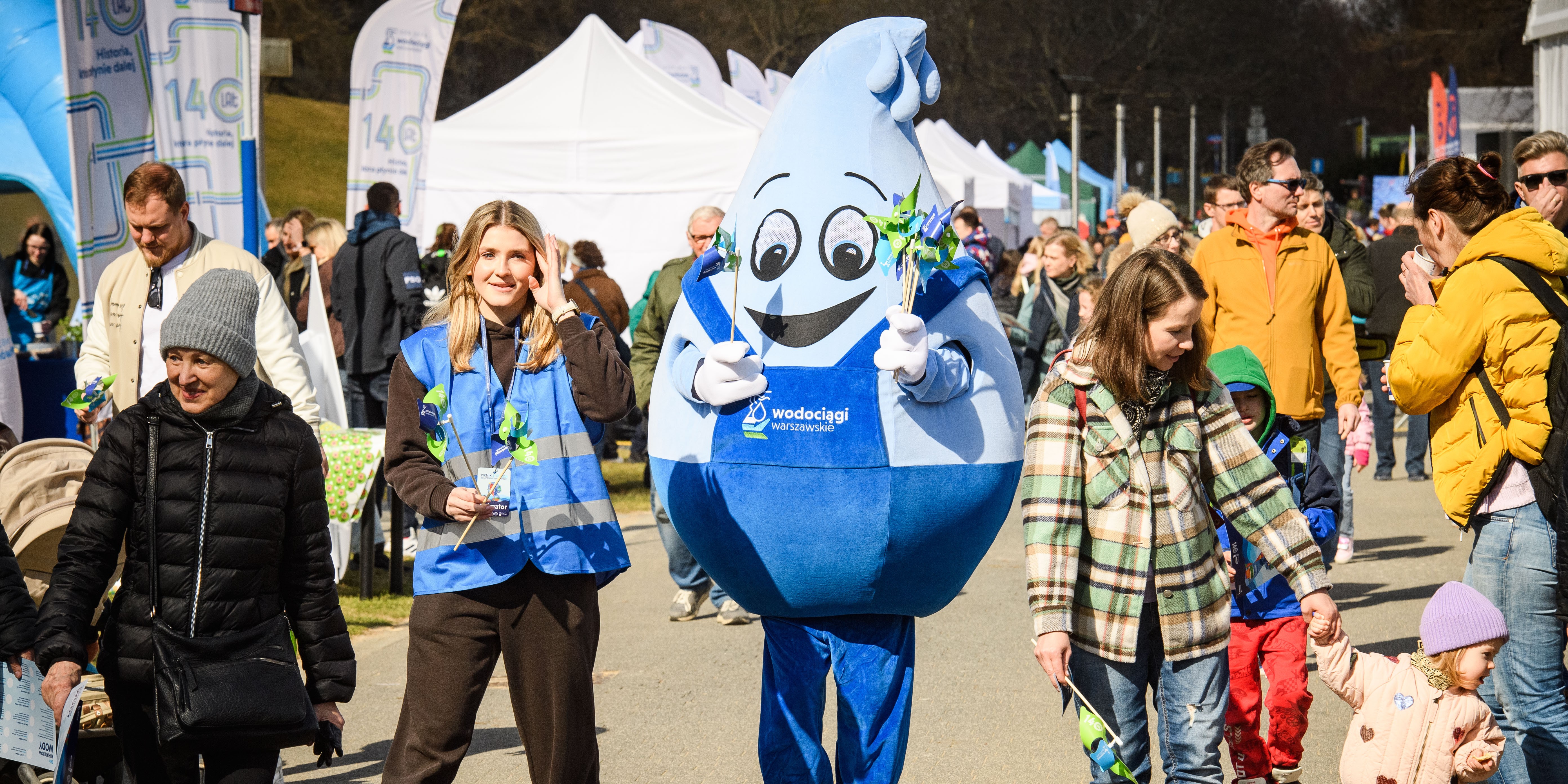 Ludzie na ulicy, pośrodku maskotka wielkości człowieka w kształcie kropli.